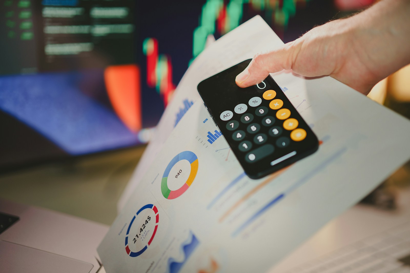 A hand using a phone calculator over printed financial charts